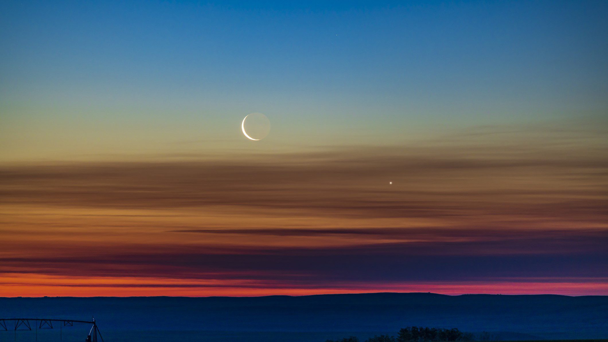 La conjunción de la media luna menguante y Venus a medida que se elevan en el cielo del amanecer del noreste en el sur de Alberta, Canadá. La luz de la Tierra se puede ver en el lado oscuro de la Luna. El cielo muestra una maravillosa transición de colores desde el naranja en el horizonte a lo largo del espectro hasta el azul en la parte superior.