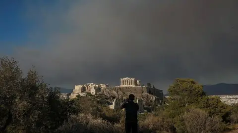 REUTERS Un hombre toma una fotografía del templo del Partenón en la cima de la colina de la Acrópolis mientras el humo de un incendio forestal en el pueblo de Varnava cubre Atenas, Grecia, el 11 de agosto de 2024.