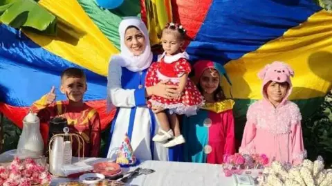 Una mujer posa con cuatro niños, todos vestidos con ropa colorida, parados frente a un mantel con los colores del arco iris y detrás de una mesa llena de comida de fiesta.